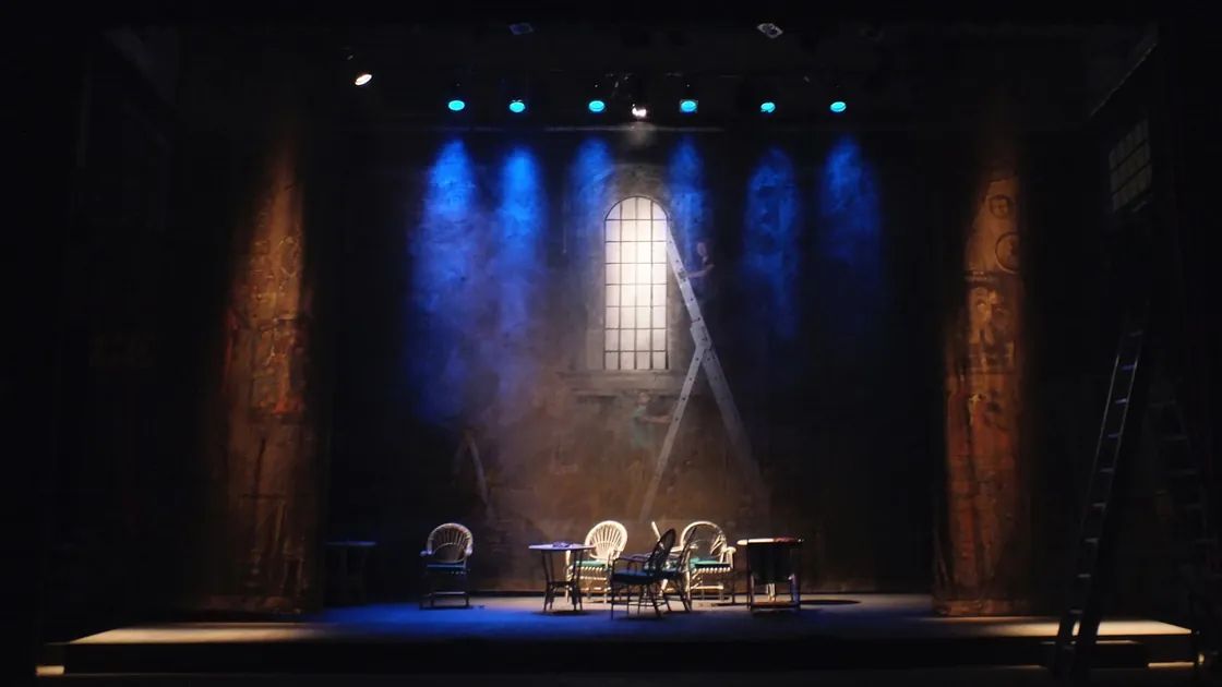 Escenario de teatro con telón de fondo pintado, mesas y sillas bajo luz azulada